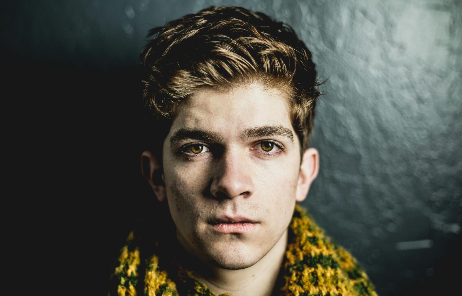 Close-up portrait of a young adult male with a serious expression against a dark textured wall.
